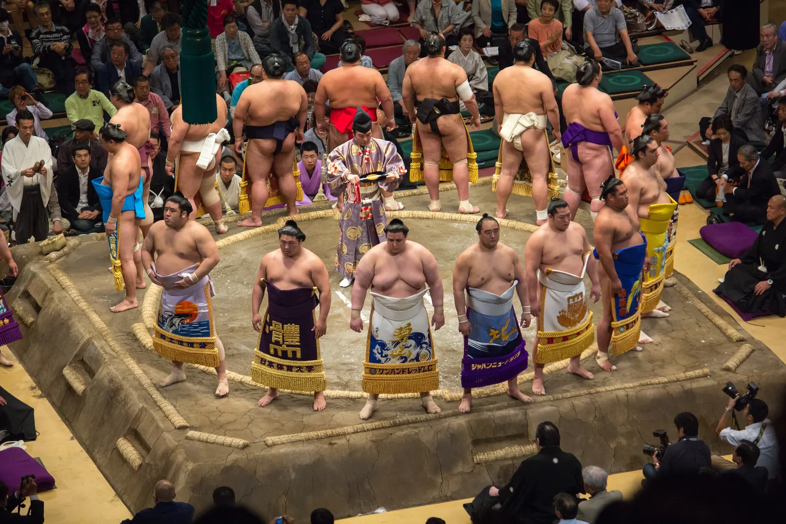 Rows of sumo wrestlers in decorated ceremonial aprons stand in a ring around the edge of a clay ring, photographed from above